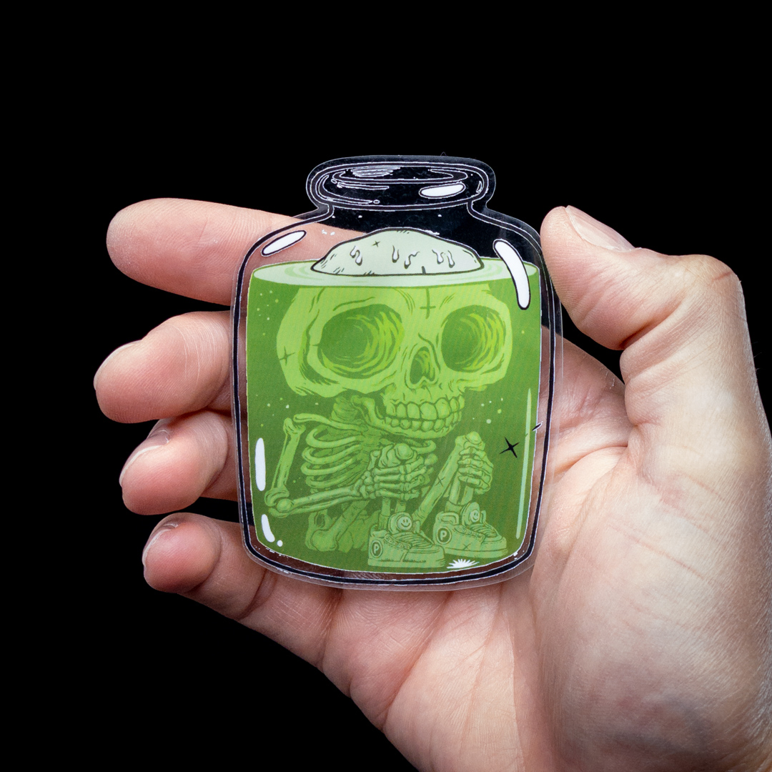 Hand holding a transparent sticker of a skeleton submerged in green liquid inside a jar, against a black background.
