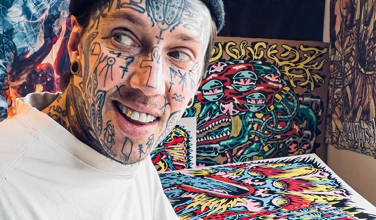 Marc UA with facial tattoos and a beanie smiles while sitting in front of his artwork.