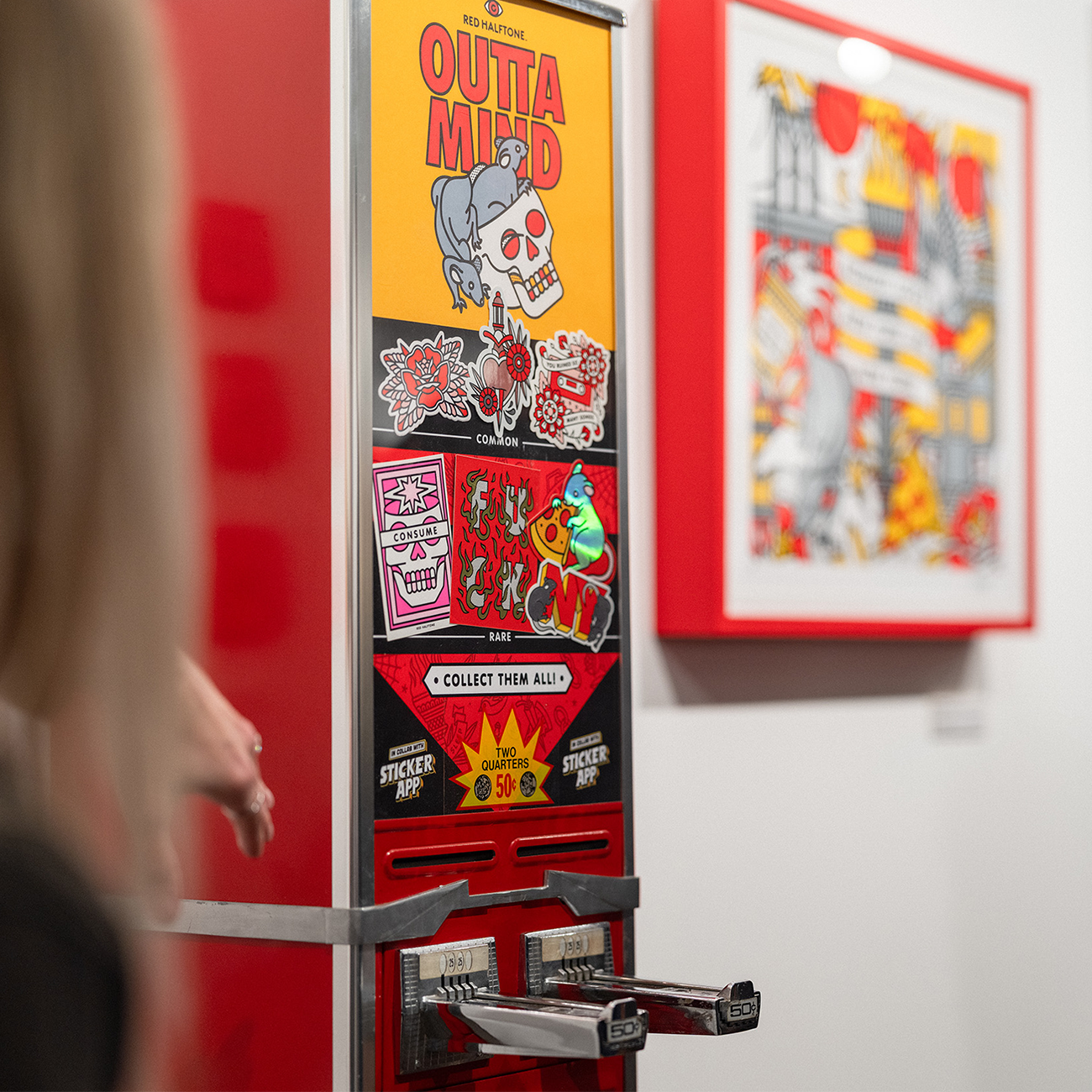 A person reaches for a vintage sticker vending machine with colorful stickers and "Outta My Mind" text, next to a framed illustration on the wall.