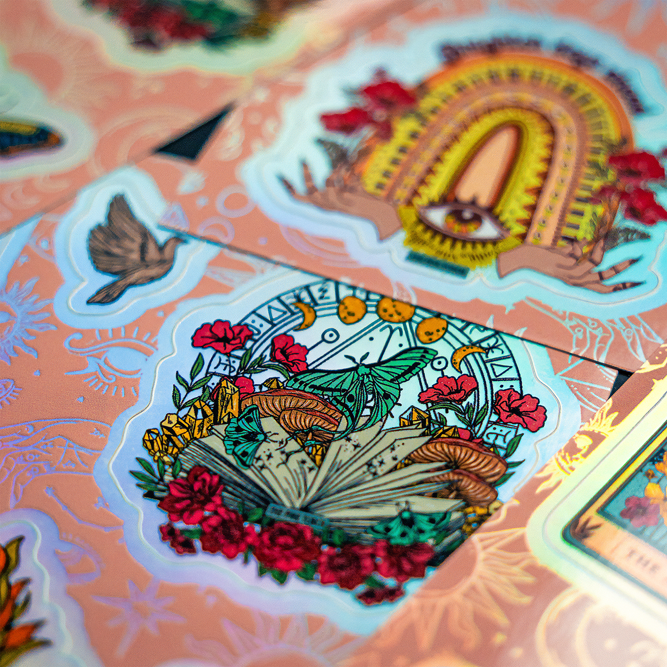 Holographic sticker sheet with a book surrounded by flowers and insects, and an geometric eye design.