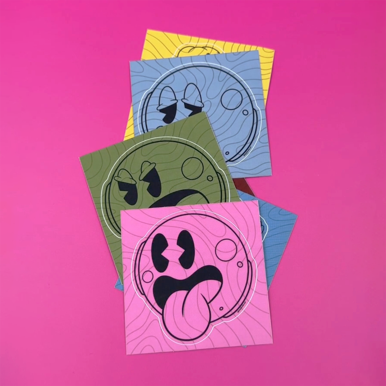 Kiss-cut Sticker smiley faces in different colours and facial expressions printed on matte vinyl, against a pink background.