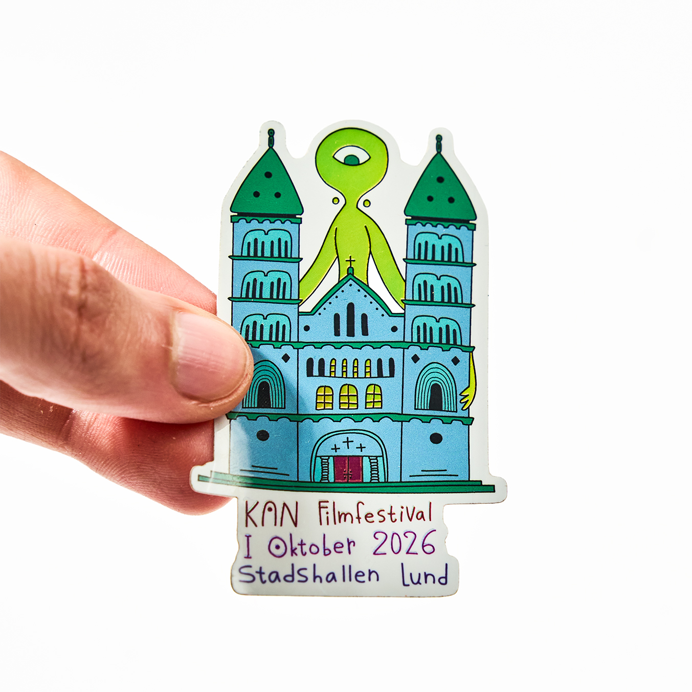 Hand holding a die-cut sticker for KAN Filmfestival, October 2026 at Stadshallen Lund, with a cathedral and a green one-eyed alien