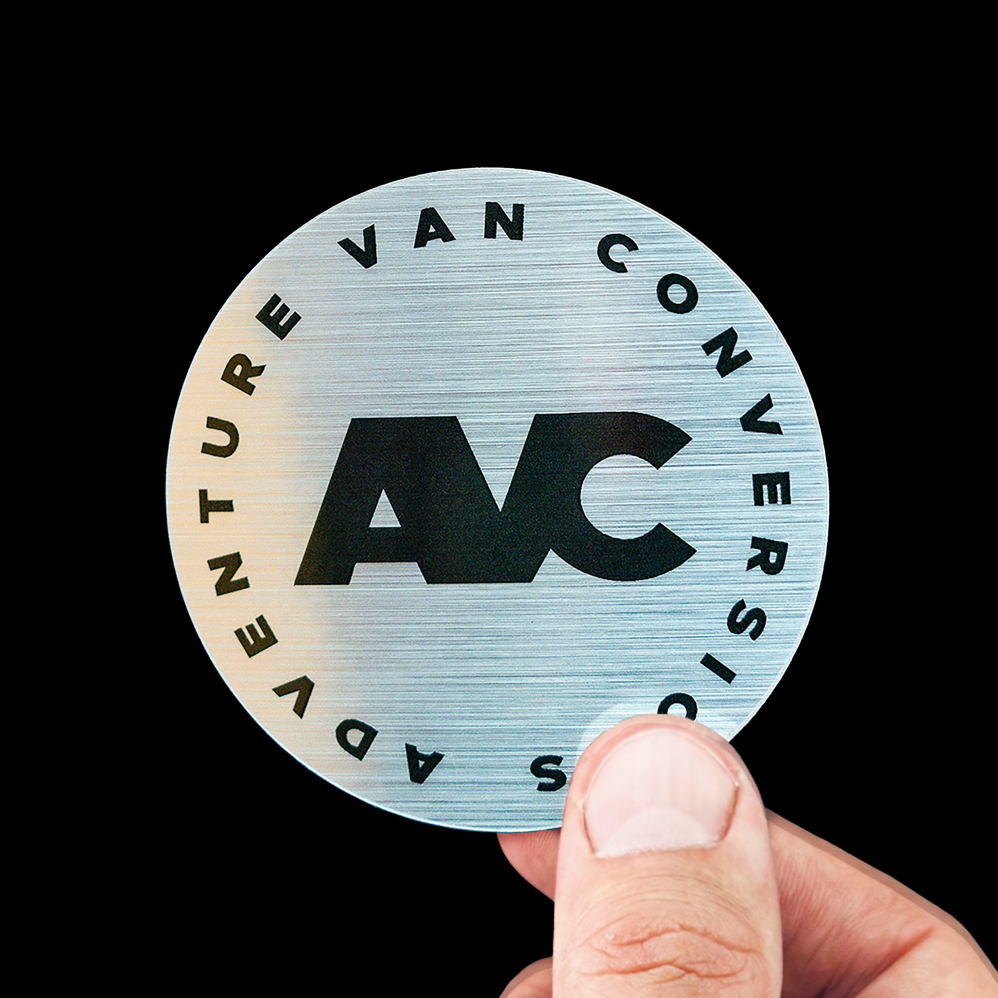 A hand holds a circular brushed aluminum sticker with "AVC" in bold letters, surrounded by "Adventure Van Conversions" text on a metallic background.