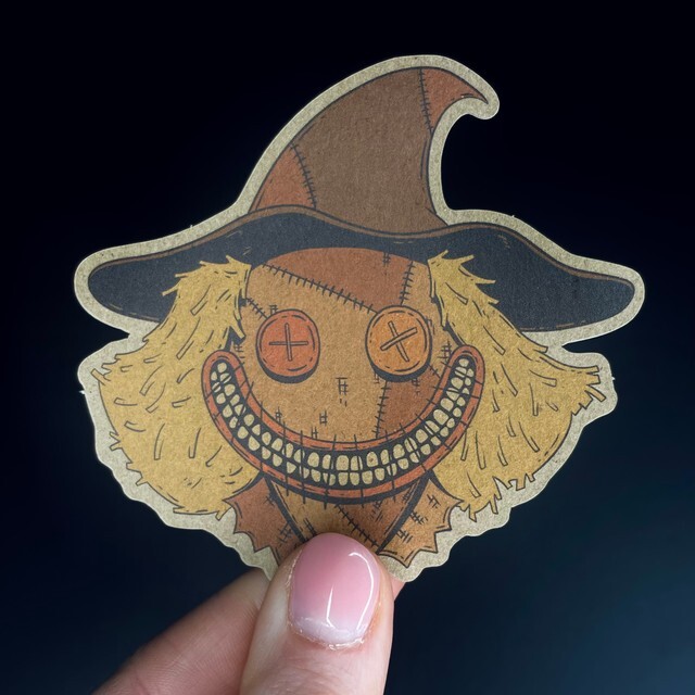 A hand holding a kraft paper sticker of a scarecrow